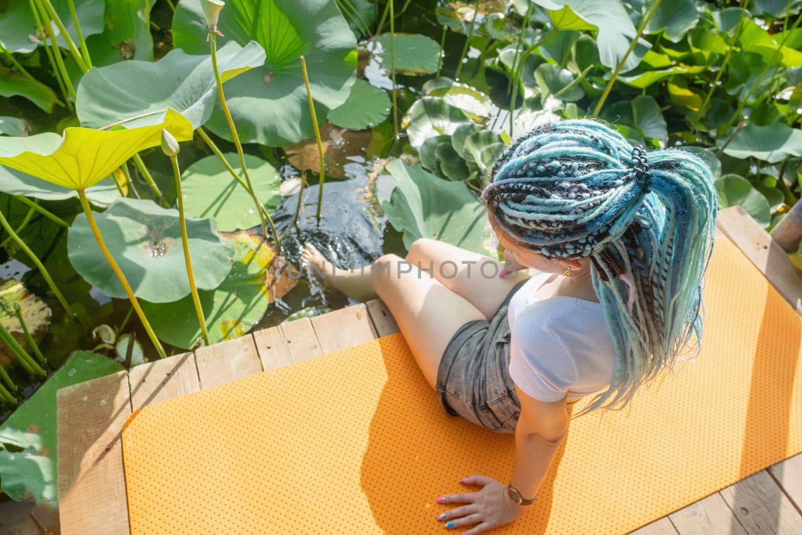 young beautiful woman with blue afro locks resting on yoga mat on wooden pierce on lotus lake enjoying nature, legs in water