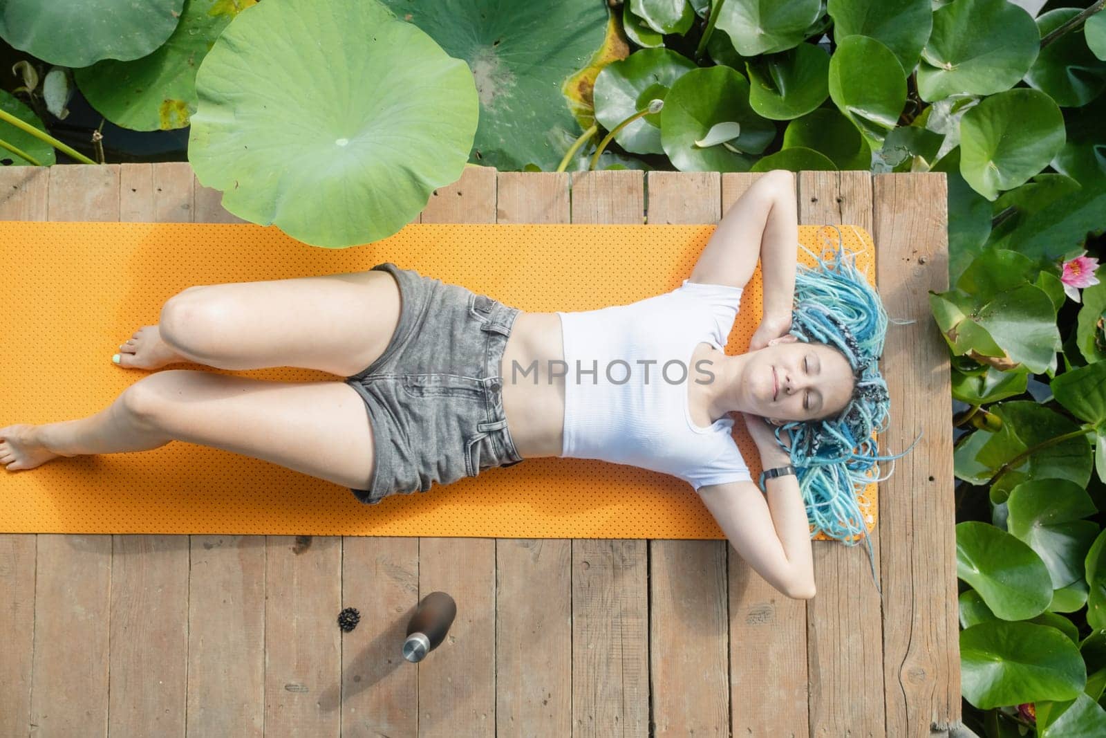 young beautiful woman with blue afro locks resting on yoga mat on wooden pierce on lotus lake enjoying nature, top view