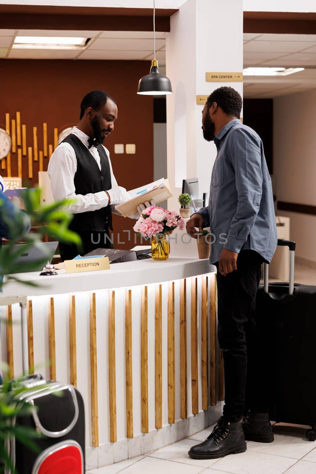 Young African American man front desk agent confirming guest reservation. Male receptionist standing behind reception counter talking with tourist arriving at hotel, checking room availability