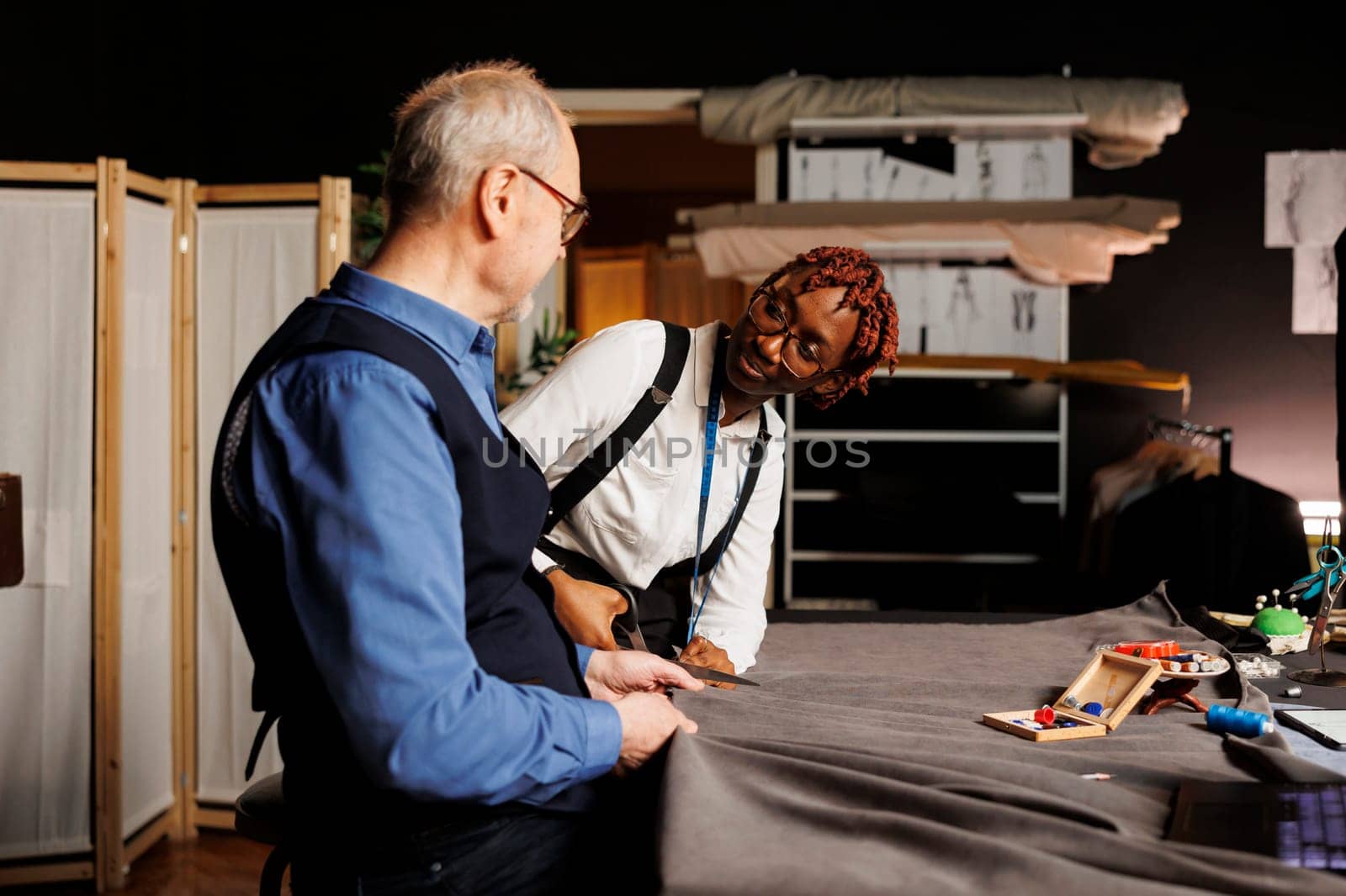 Helpful old master dressmaker guiding happy apprentice with cutting textile material for upcoming bespoken sartorial collection. Couturiers in tailoring studio working on making stylish fashion attire