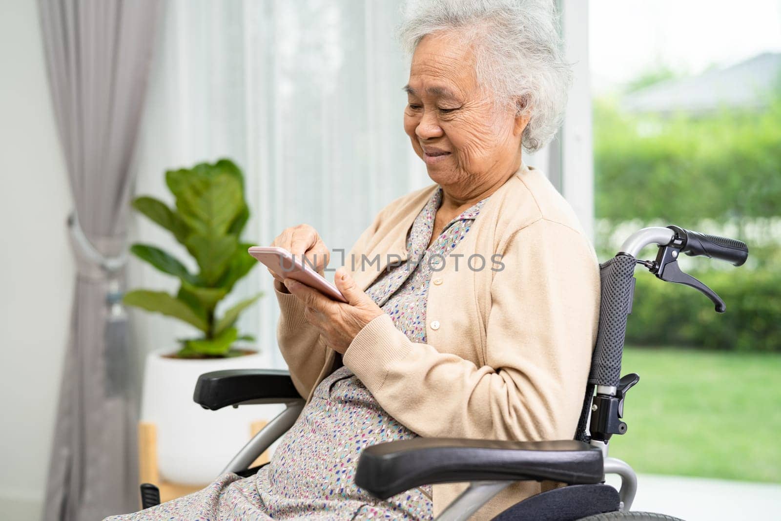 Asian elderly woman patient use mobile phone while sitting and happy on wheelchair at home.