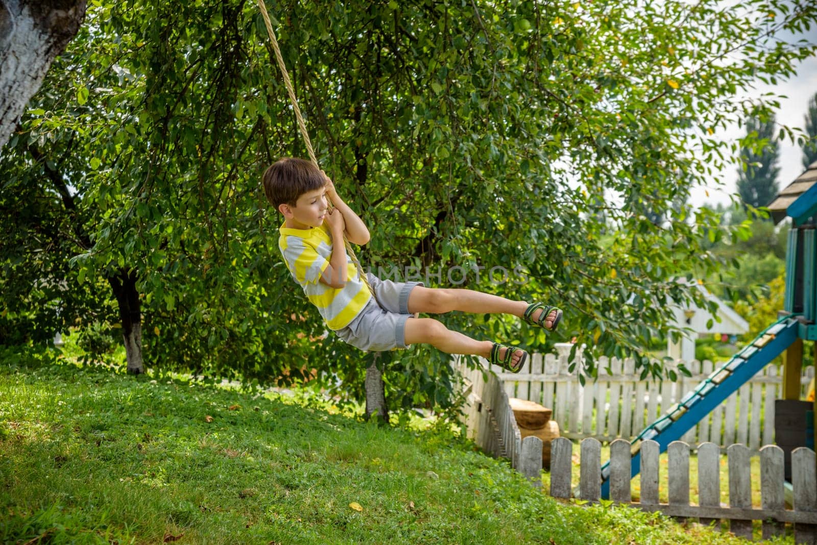 Happy little boy is having fun on a rope swing which he has found while having rest outside city. Active leisure time with children.