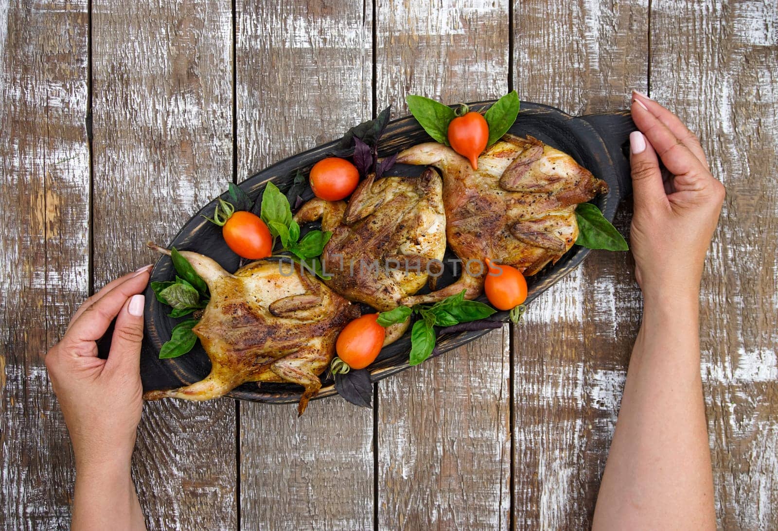 Women's hands hold a wooden dish with grilled quails. Tomatoes and bell peppers are lying on a wooden table.