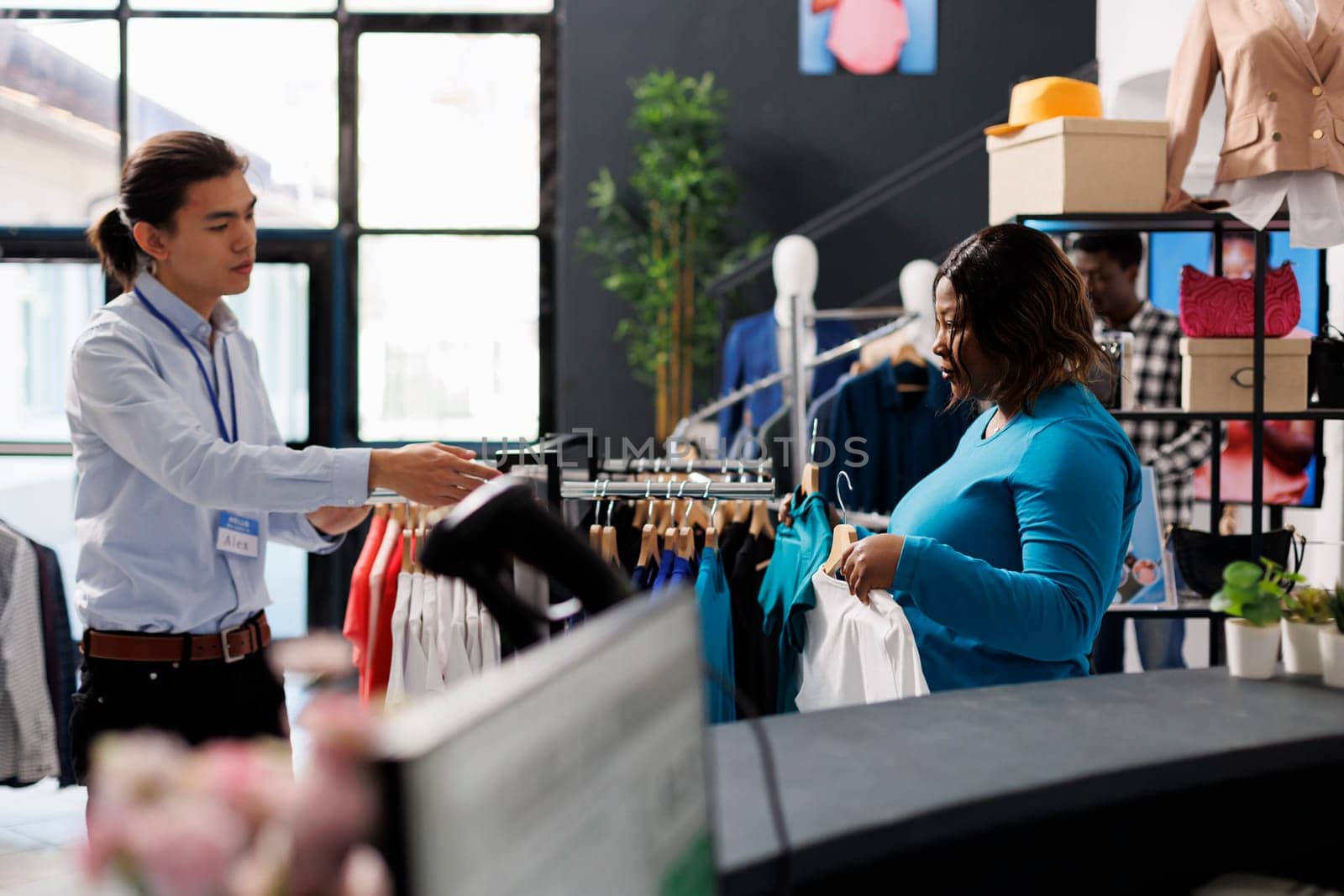 Asian employee discussing with stylish client, showing trendy clothes in modern boutique. African american customer shopping for casual wear, looking at stylish merchandise in clothing store