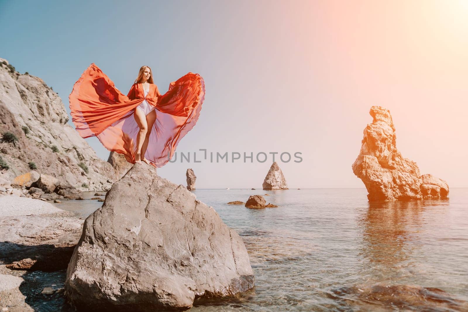 Woman travel sea. Young Happy woman in a long red dress posing on a beach near the sea on background of volcanic rocks, like in Iceland, sharing travel adventure journey by panophotograph