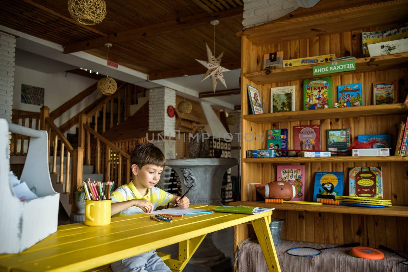 Little cutie curly haired baby boy sitting on floor of eco living room interior next to books shelf and drawing with colorful felt tip pens in album. Creative hobby for kids and playtime activity.