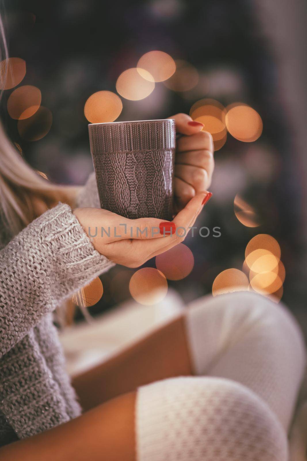 Close-up of a cute young woman sitting in the bed and holding cup of tea or coffee, surrounded with Christmas lights. 