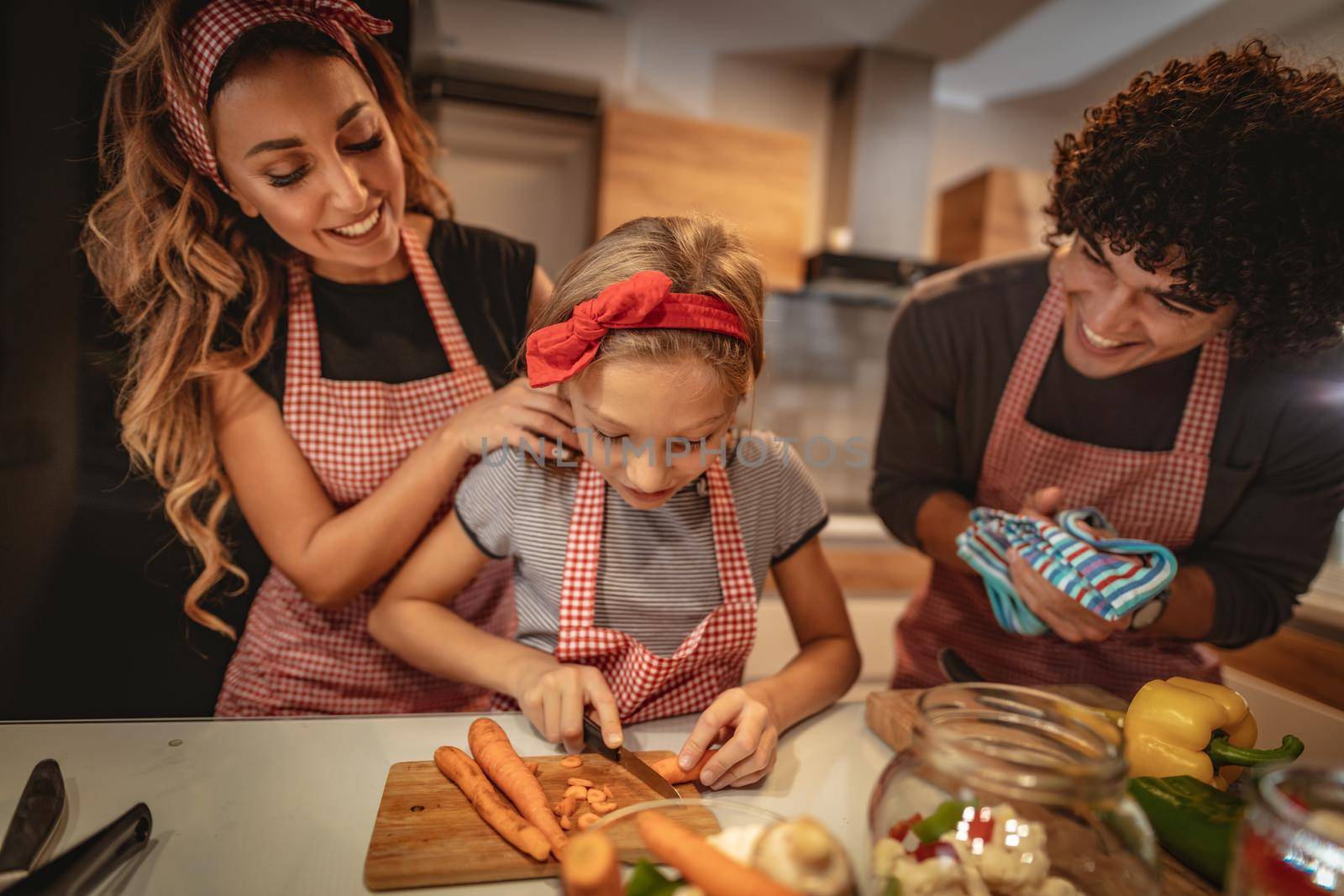 Happy parents and their daughter cooking together in the kitchen while little girl trying to cut carrot.