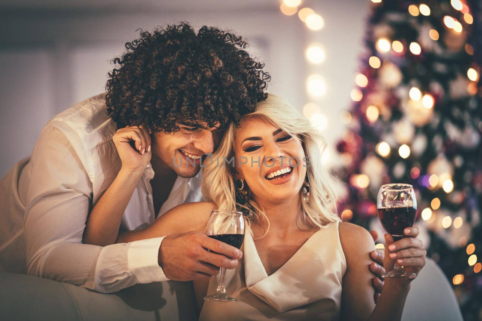 Young couple celebrating Christmas at home. They are relaxing with glass of red vine, covered with blanket on a sofa in an apartment.