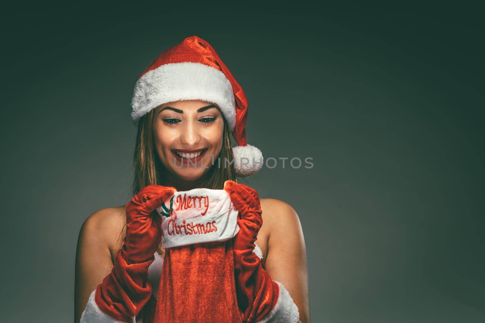 Cheerful beautiful young woman in Santa Claus costume enjoying and surprisingly looking what is inside of a red christmas stocking.