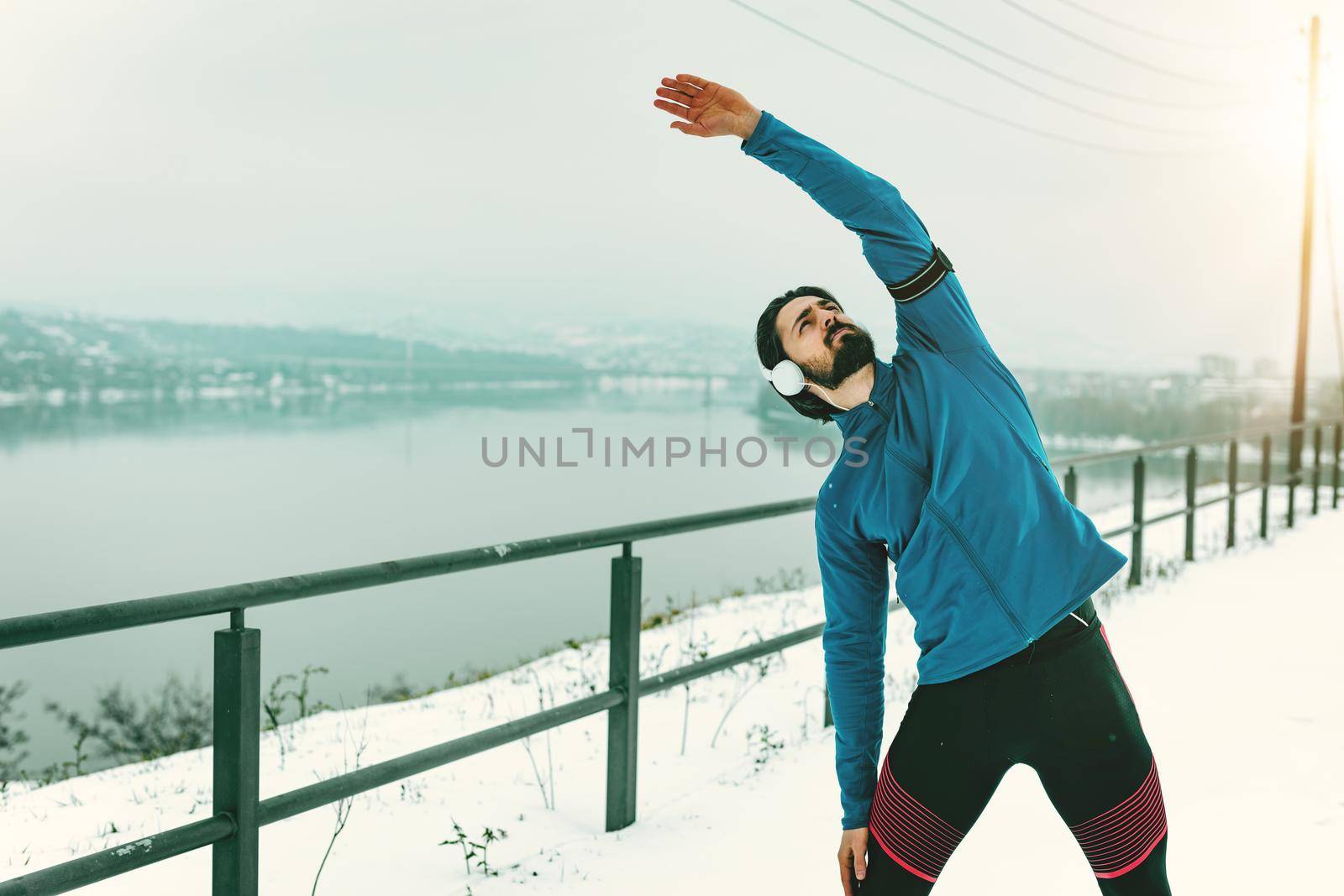 Active young man with headphones on his ears stretching and doing exercises in the public place during the winter training outside.