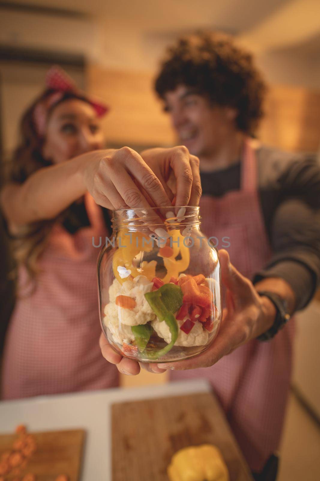 Cute couple is putting vegetables in a jar and smiling while making pickle in kitchen at home