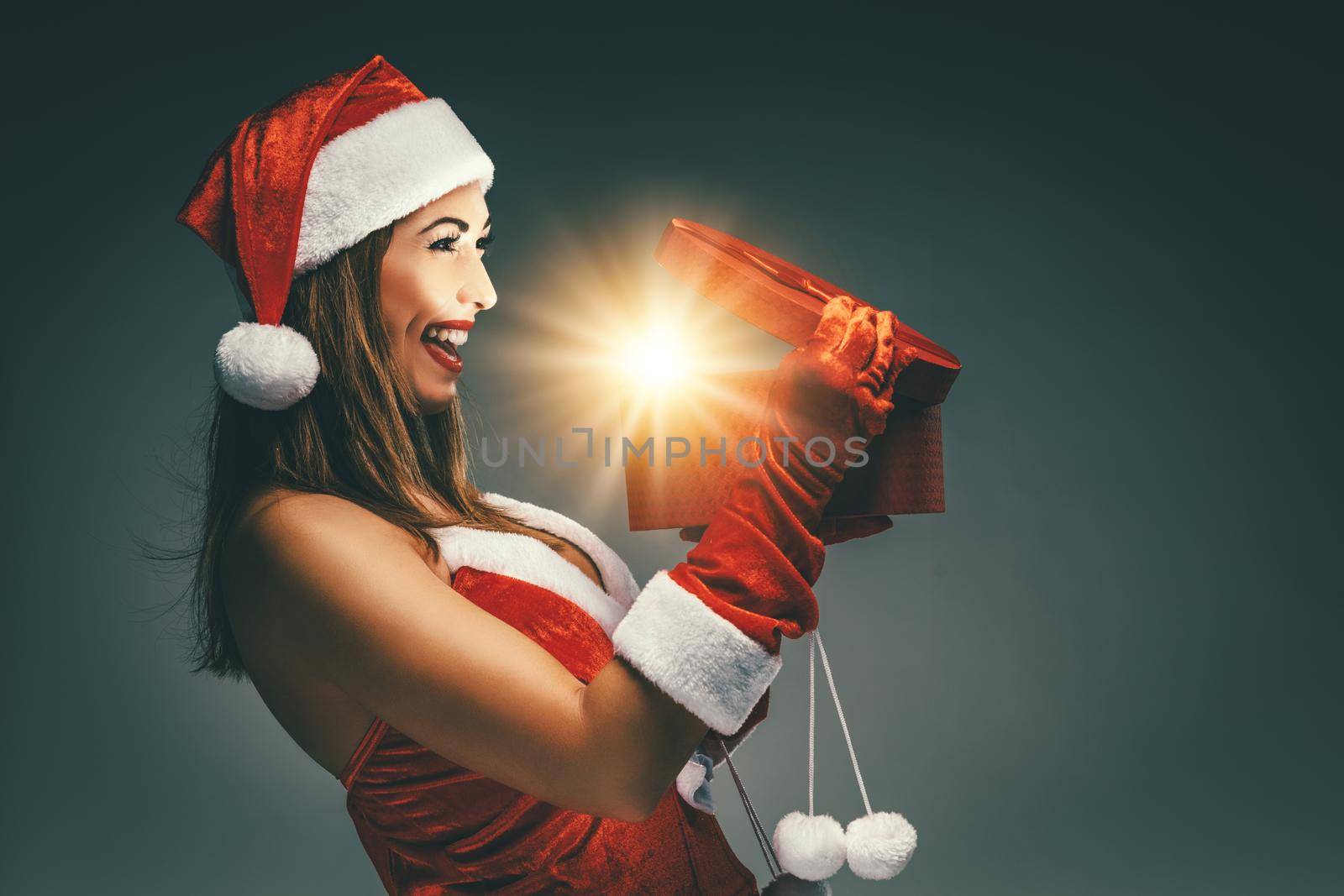 Beautiful young smiling woman in Santa Claus costume holding red present and having fun looking what is in it.