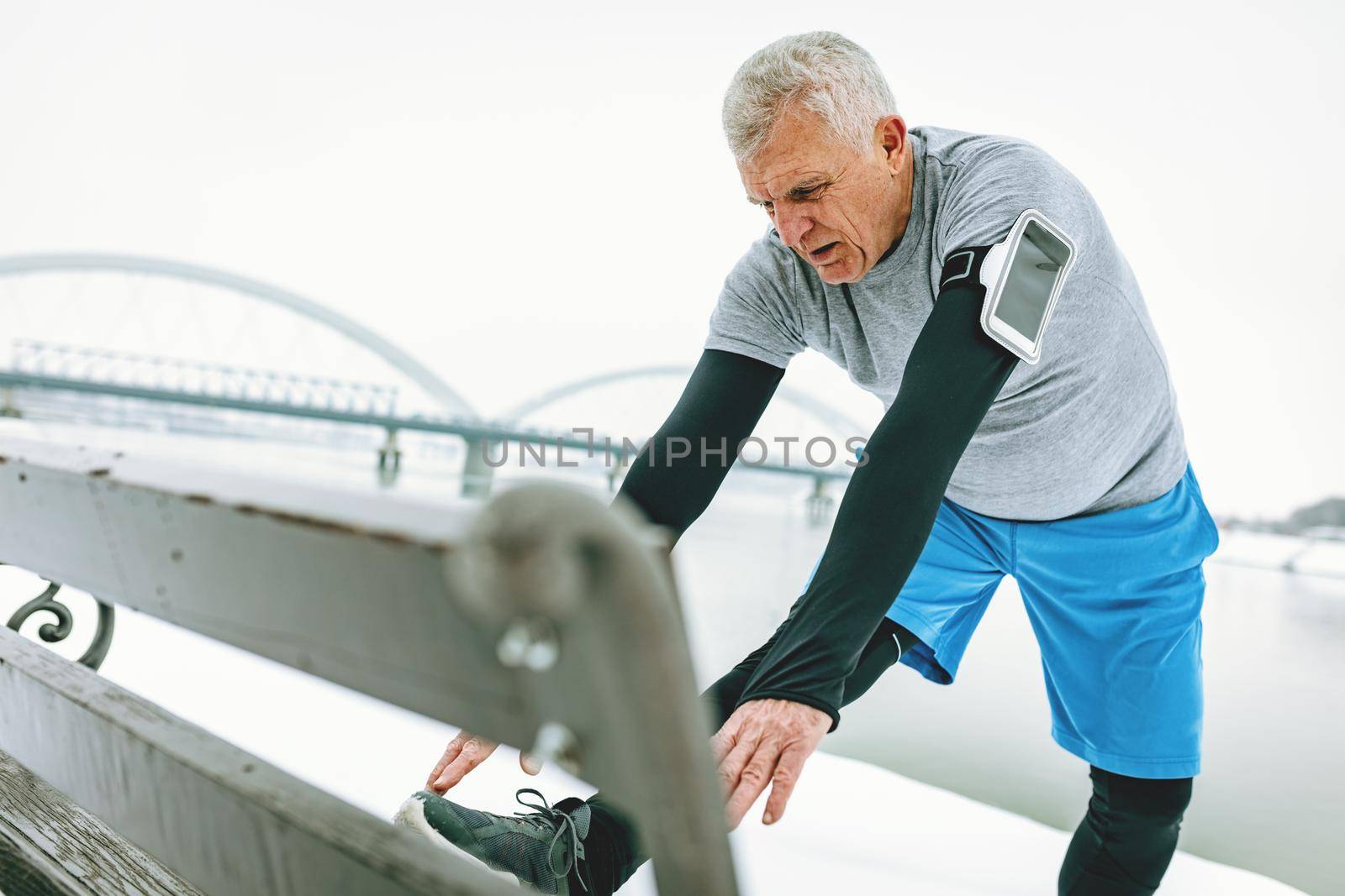 Active senior man stretching and doing exercises on the bench by the river during the winter training outside in. Copy space.