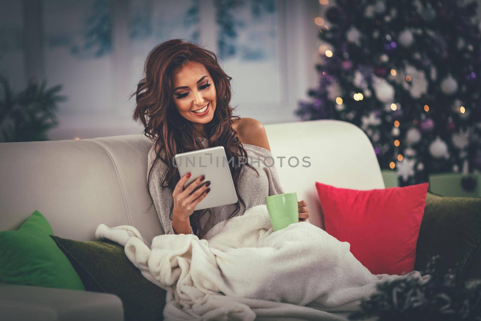 Beautiful smiling woman sitting wrapped in a blanket on Christmas eve with cup of tea and using tablet.