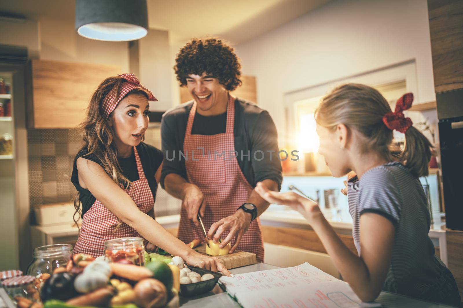 Happy parents and their daughter cooking together in the kitchen while little girl doing her homework on the kitchen table.