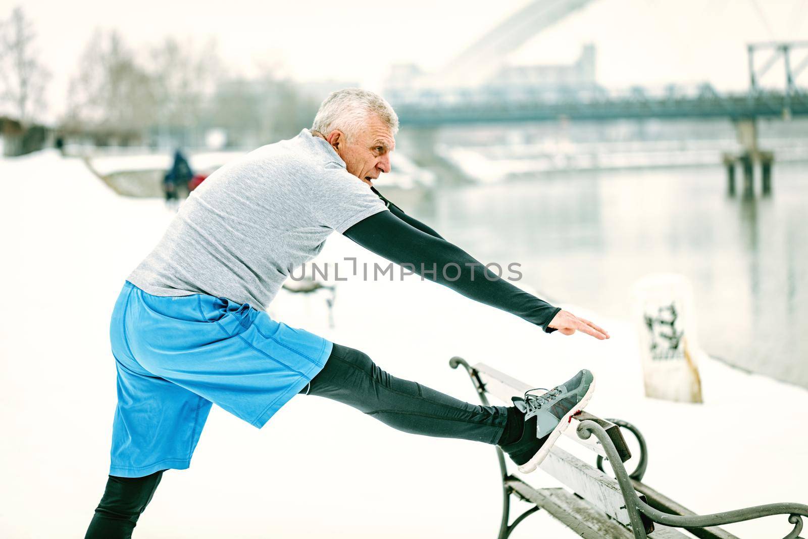 Active senior man athlete stretching leg on the bench and doing exercises by the river during the winter training outside in. Copy space.