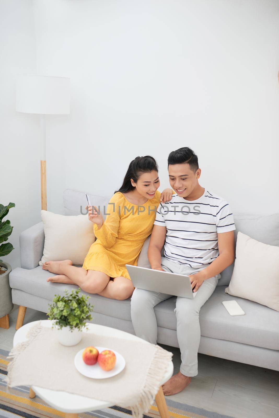 Pretty Asian woman and handsome man relaxing on couch with laptop and surfing Internet while making purchases online by makidotvn