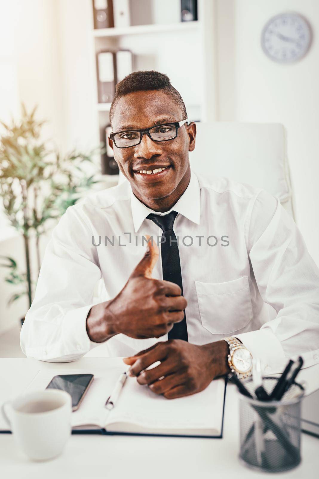 African businessman is sitting in the office, satisfied with success and shows thumb up.