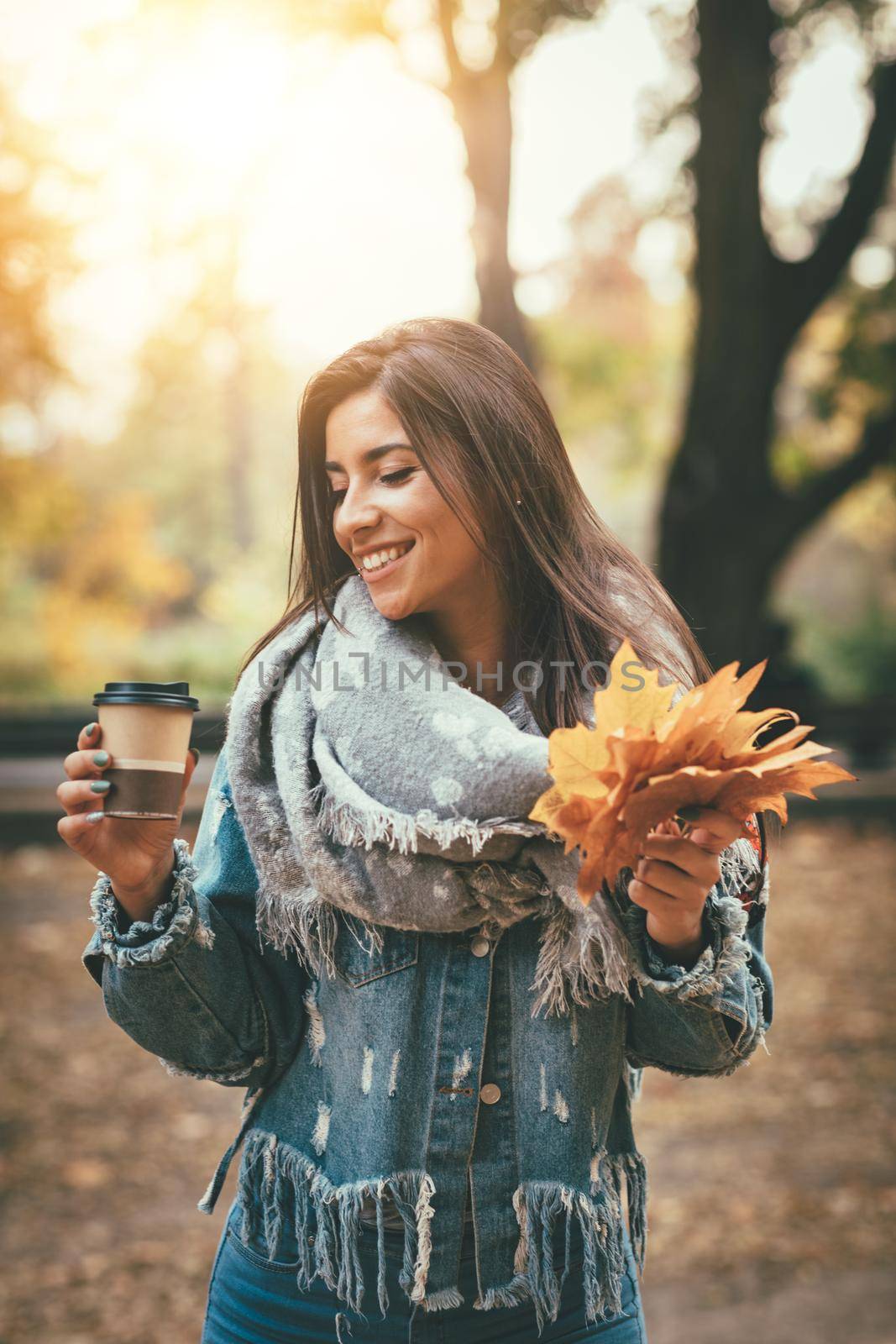Cute young woman enjoying in sunny forest in autumn colors. She is holding golden yellow leaves and cup of cofee.
