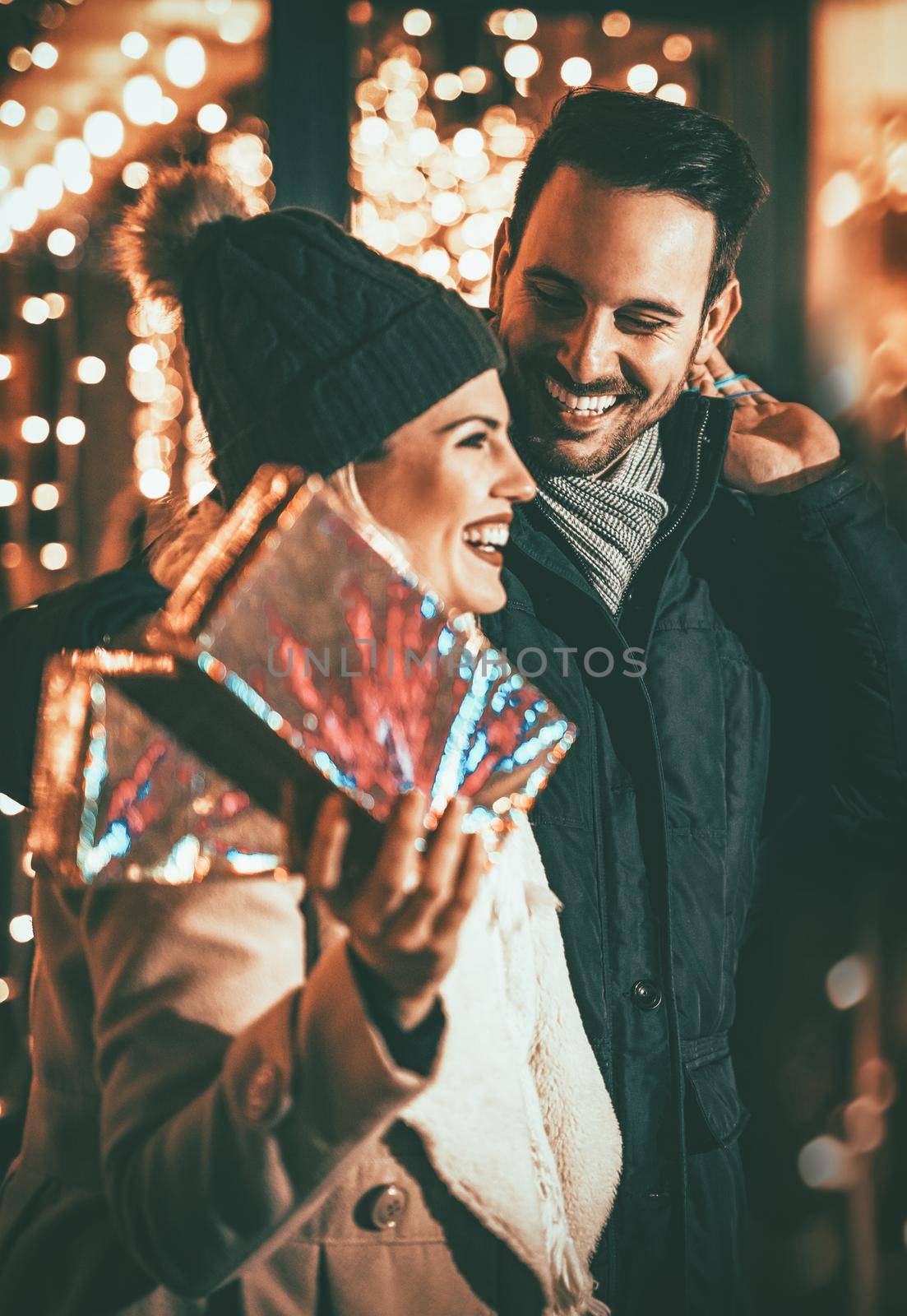 Young happy smiling couple in shopping passes in front of shopping mall. Girl is carrying gift boxes in her hand.
