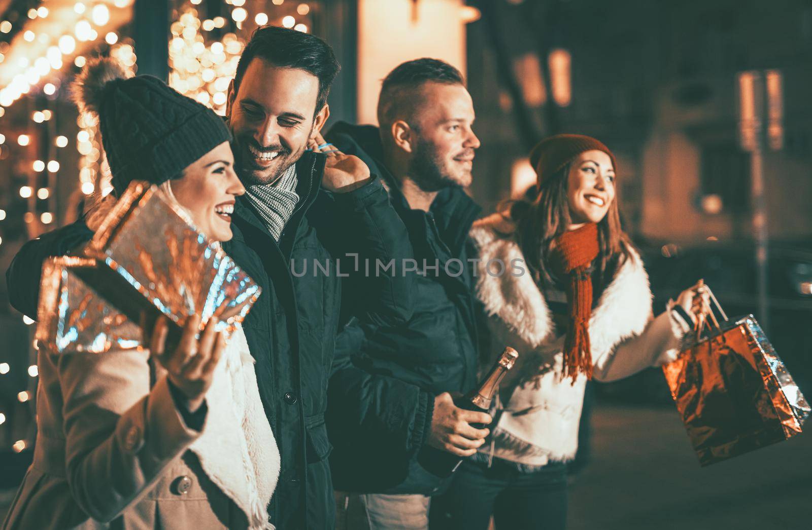 Four smiling young friends standing outside and holding many shopping paper bags and boxes in city holiday night. Selective focus. Focus on foreground.