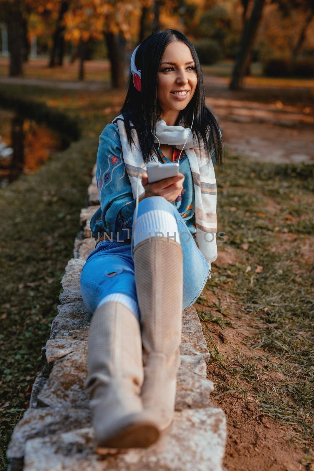 Young woman listening music on a smartphone with headphones on her ears and sitting in a park in a wonderful autumn day.