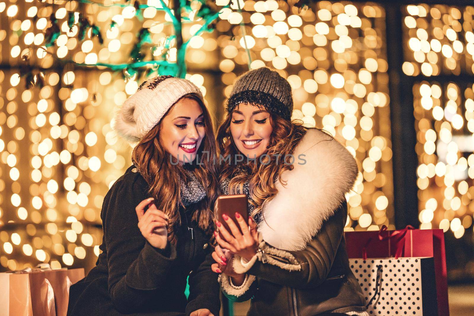 Cheerful young female friends are having fun in city street at night. They are surfing on social network at smartphone and talking something funny.