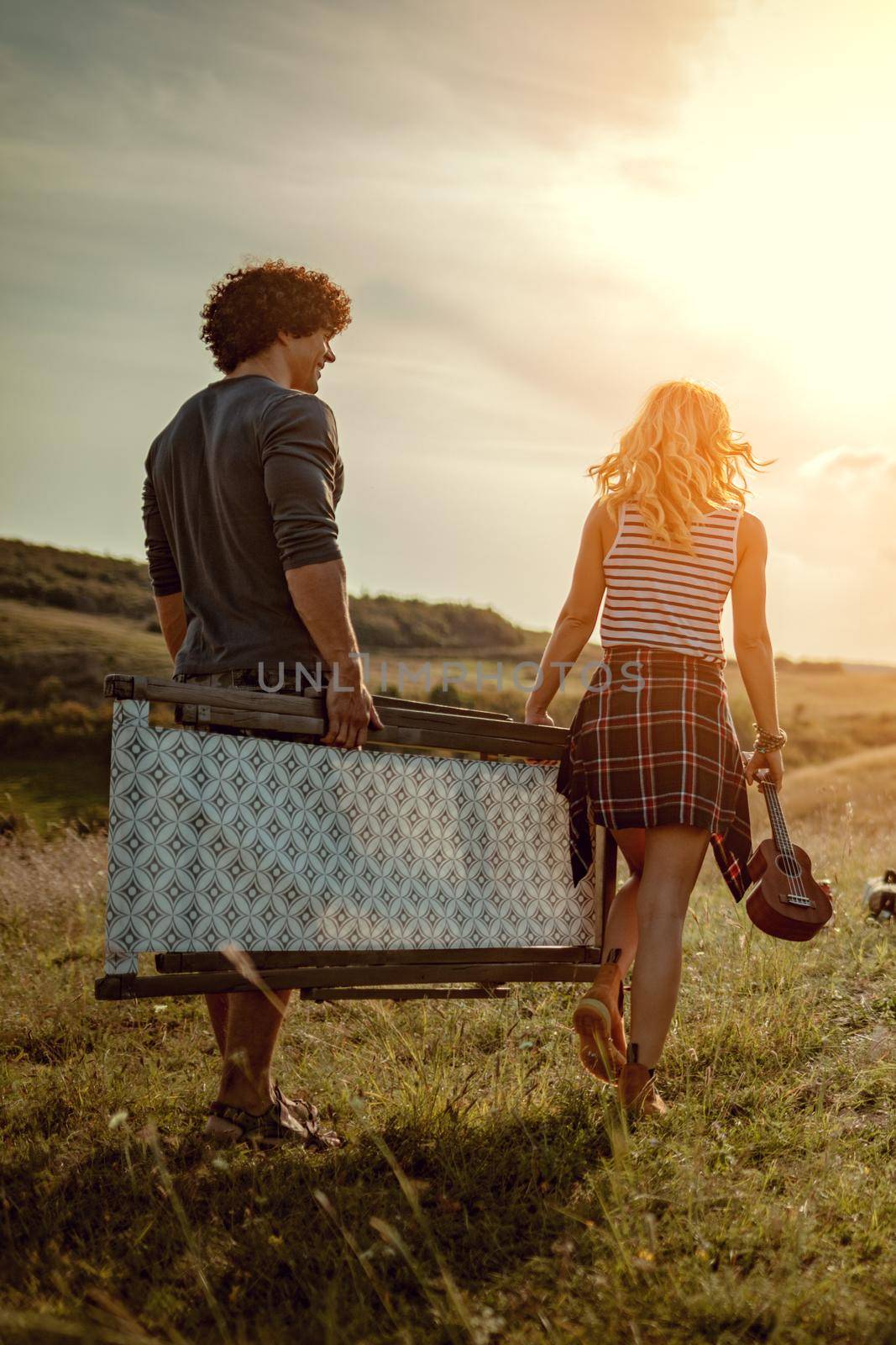 The young couple is preparing for camping. They're holding a resting chair and searching for a suitable place in a sunset.