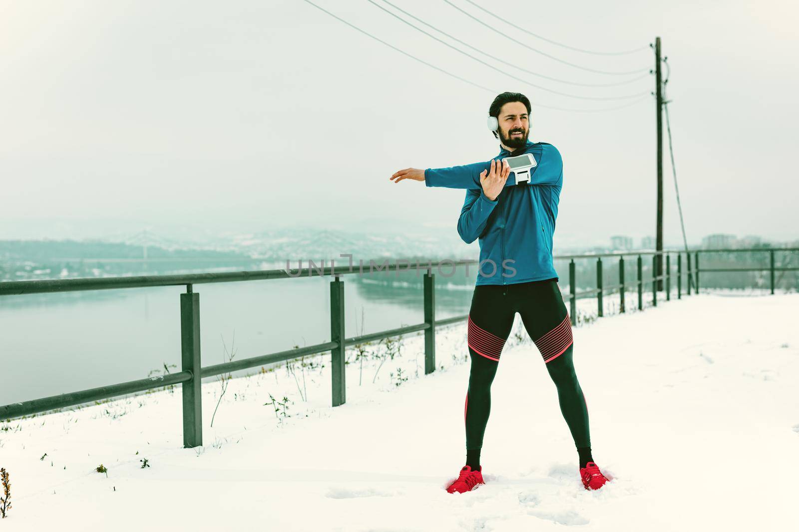 Active young man with headphones on his ears stretching and doing exercises in the public place during the winter training outside beside the river.