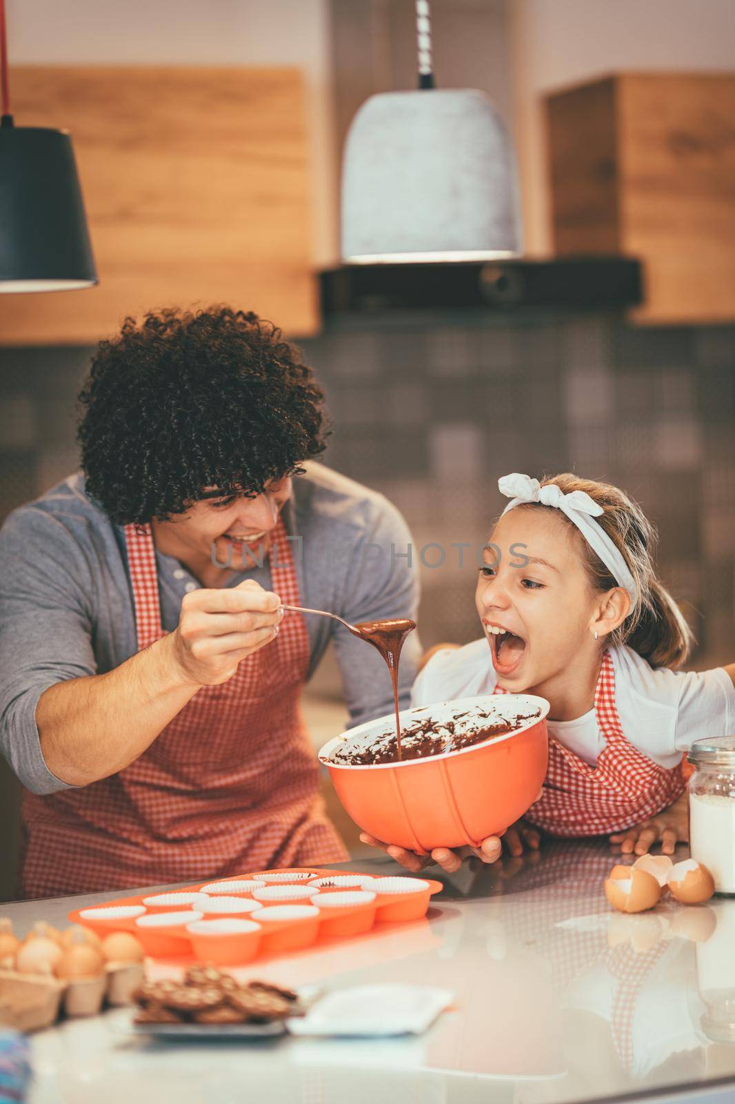 Happy father and his daughter are preparing cookies together in the kitchen. Father is pouring chocolate dough into molds and little girl wants to eat it.