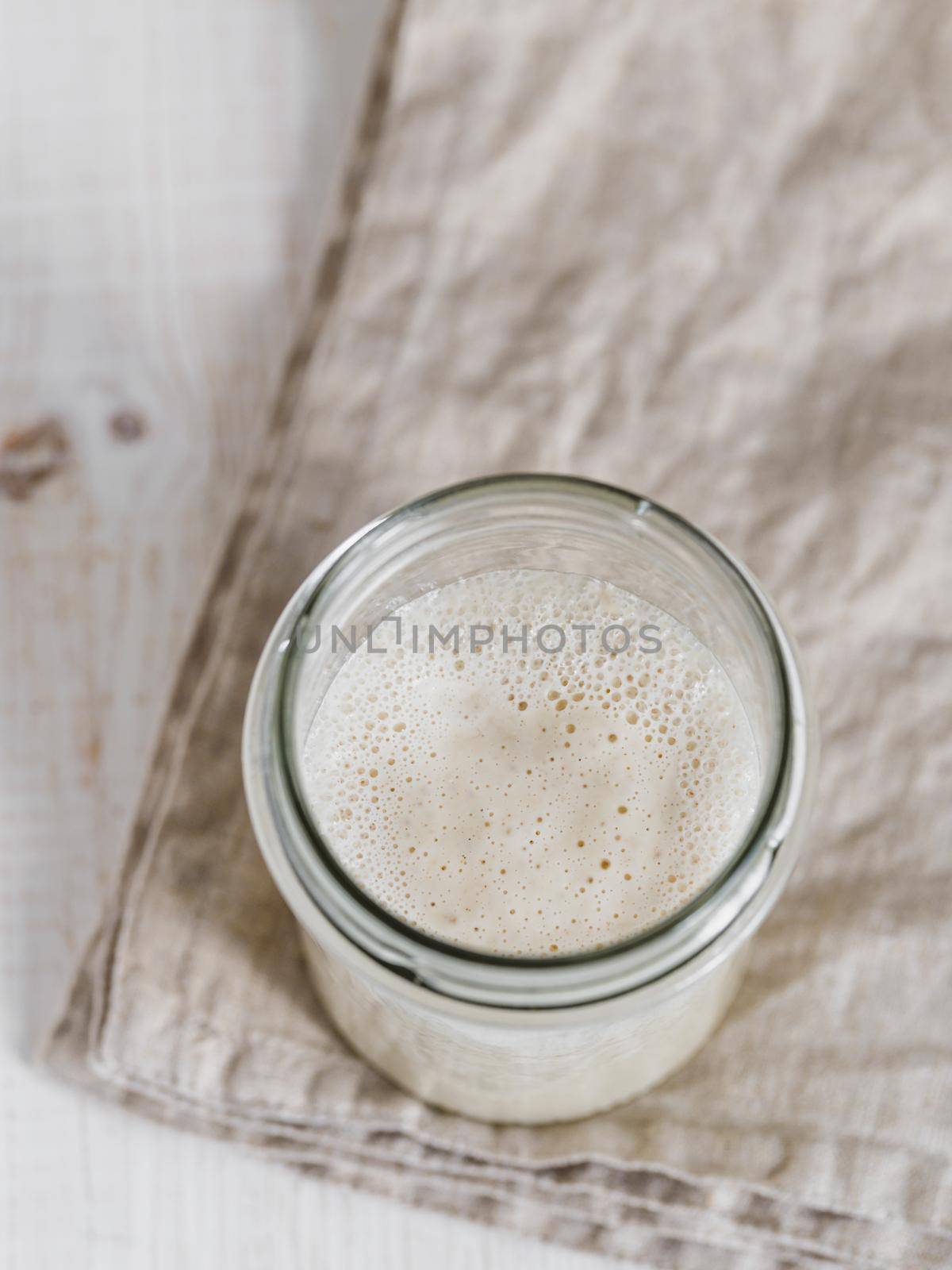 Wheat sourdough starter. Top view of glass jar with sourdough starter on white wooden background. Copy space for text or design. Vertical.