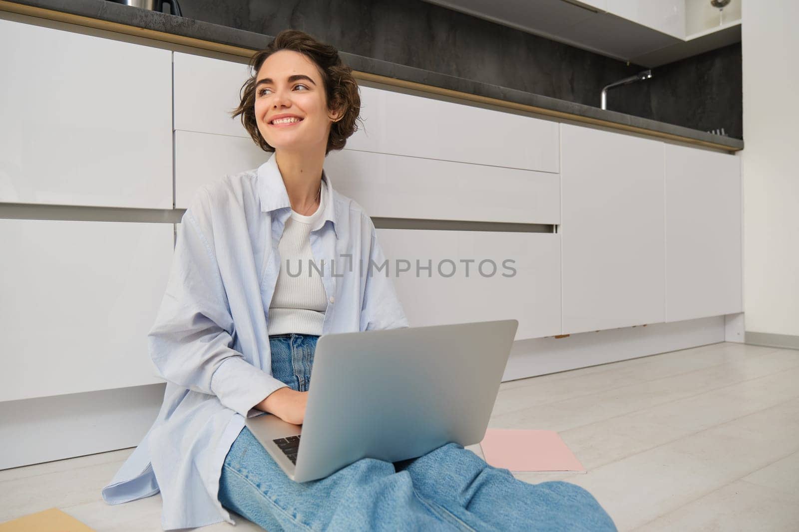 Young woman works from home on kitchen floor, does homework on laptop, using internet on computer, freelancing.