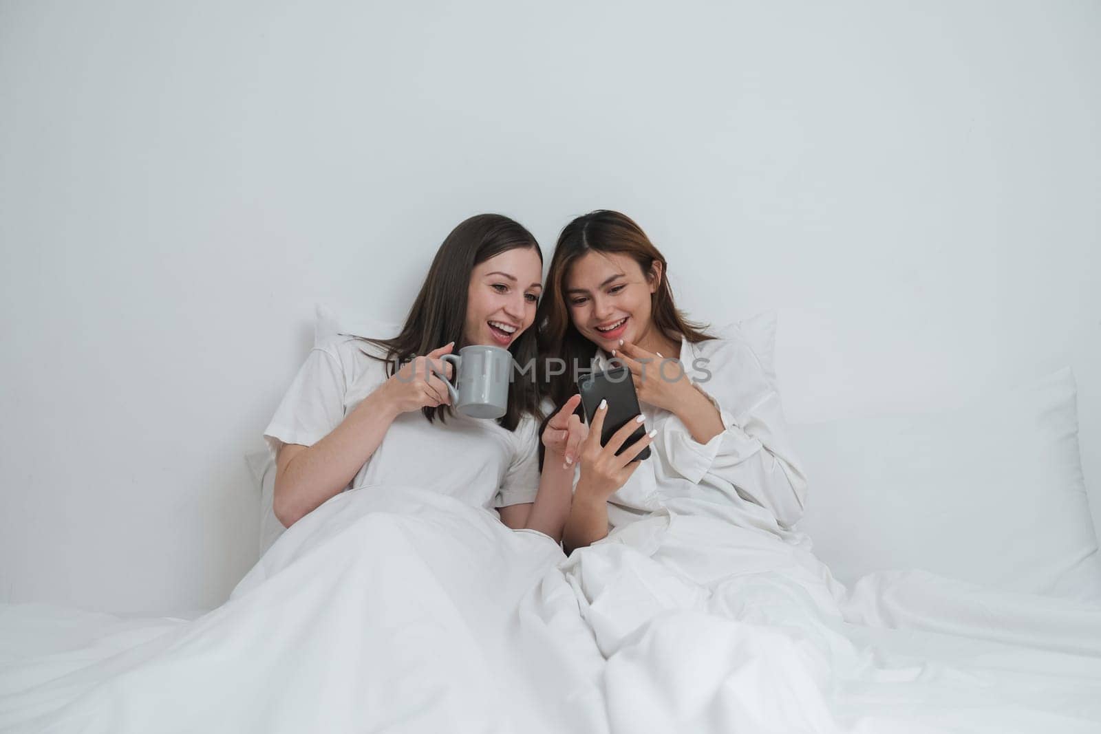 Two Asian cheerful happy female girlfriends having a coffee sitting on the bed while looking at the smartphone. Concept lifestyle lesbian couple by nateemee