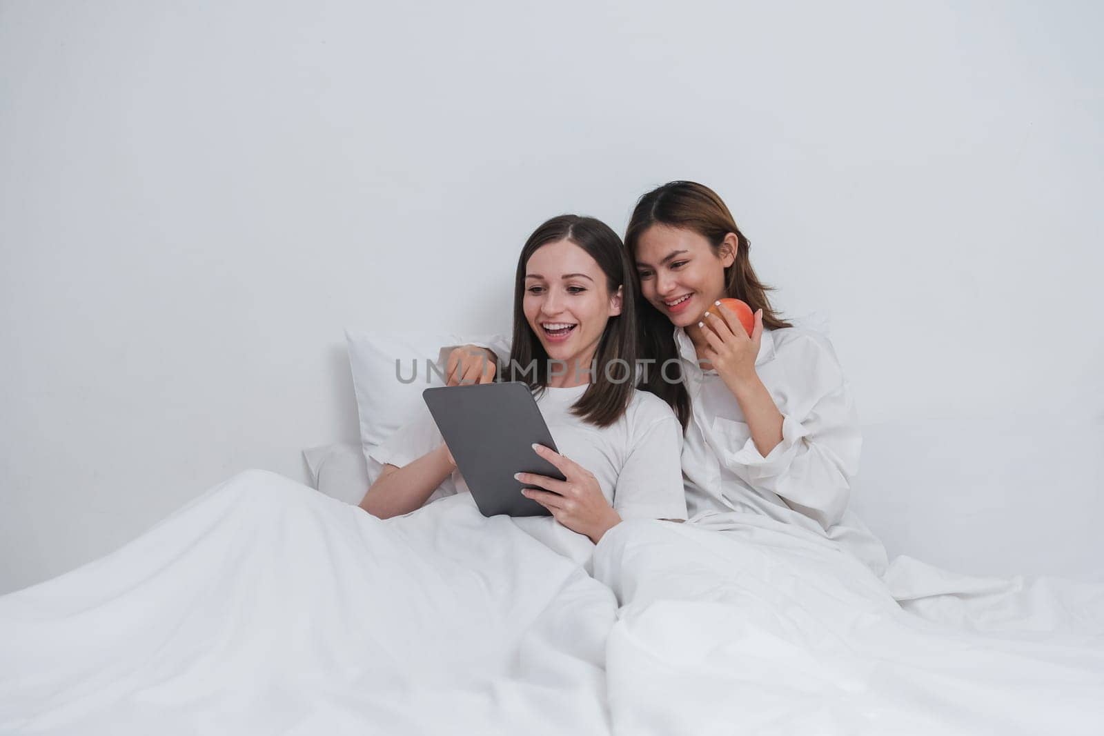 LBGT Couple of cute lesbian women waking up in the morning on white bed in bedroom while laughing and looking at tablet together.