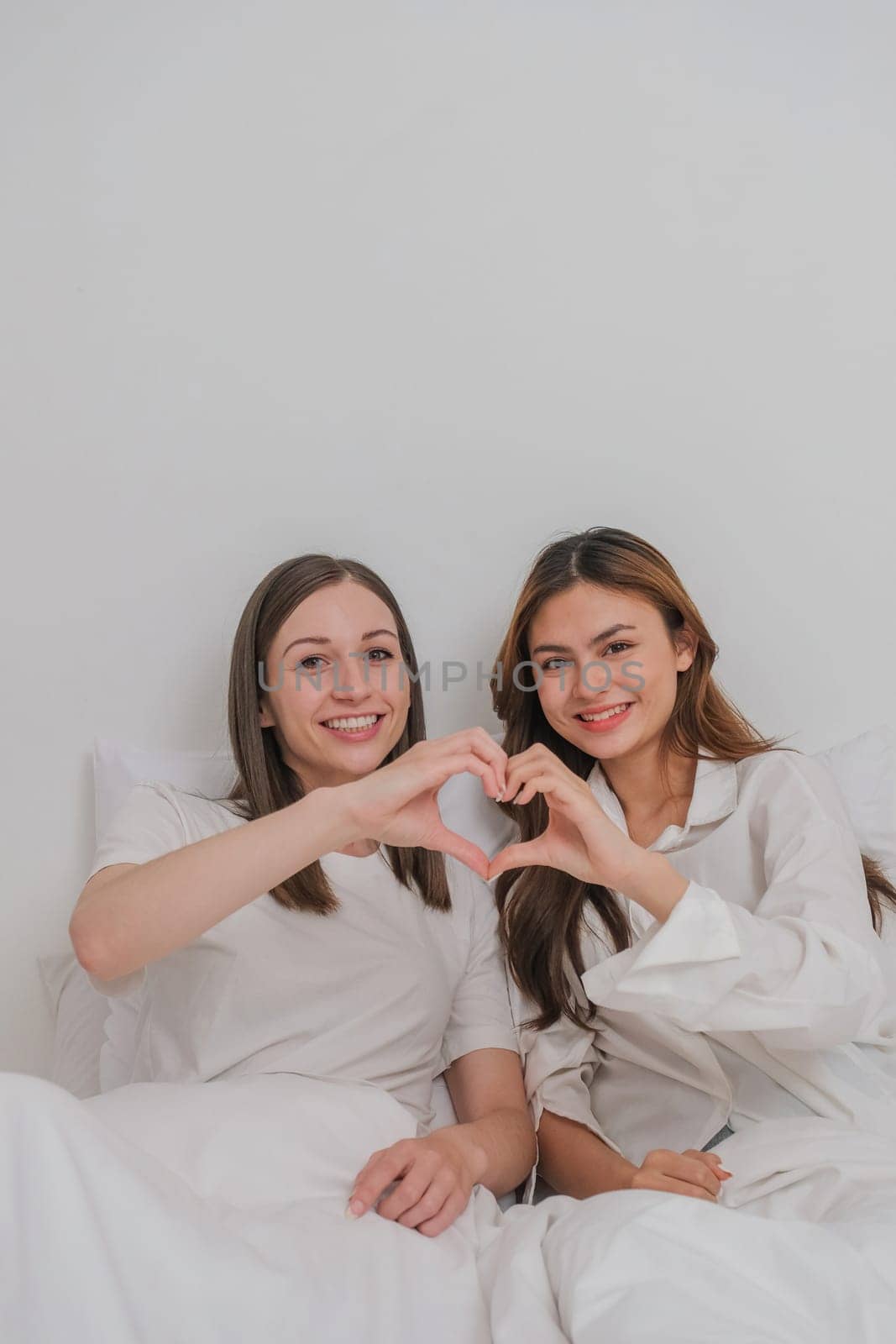 Lesbian couple together concept. Portrait of a smiling young lesbian couple other while sitting together and hand made heart symbol in the morning at bedroom by nateemee