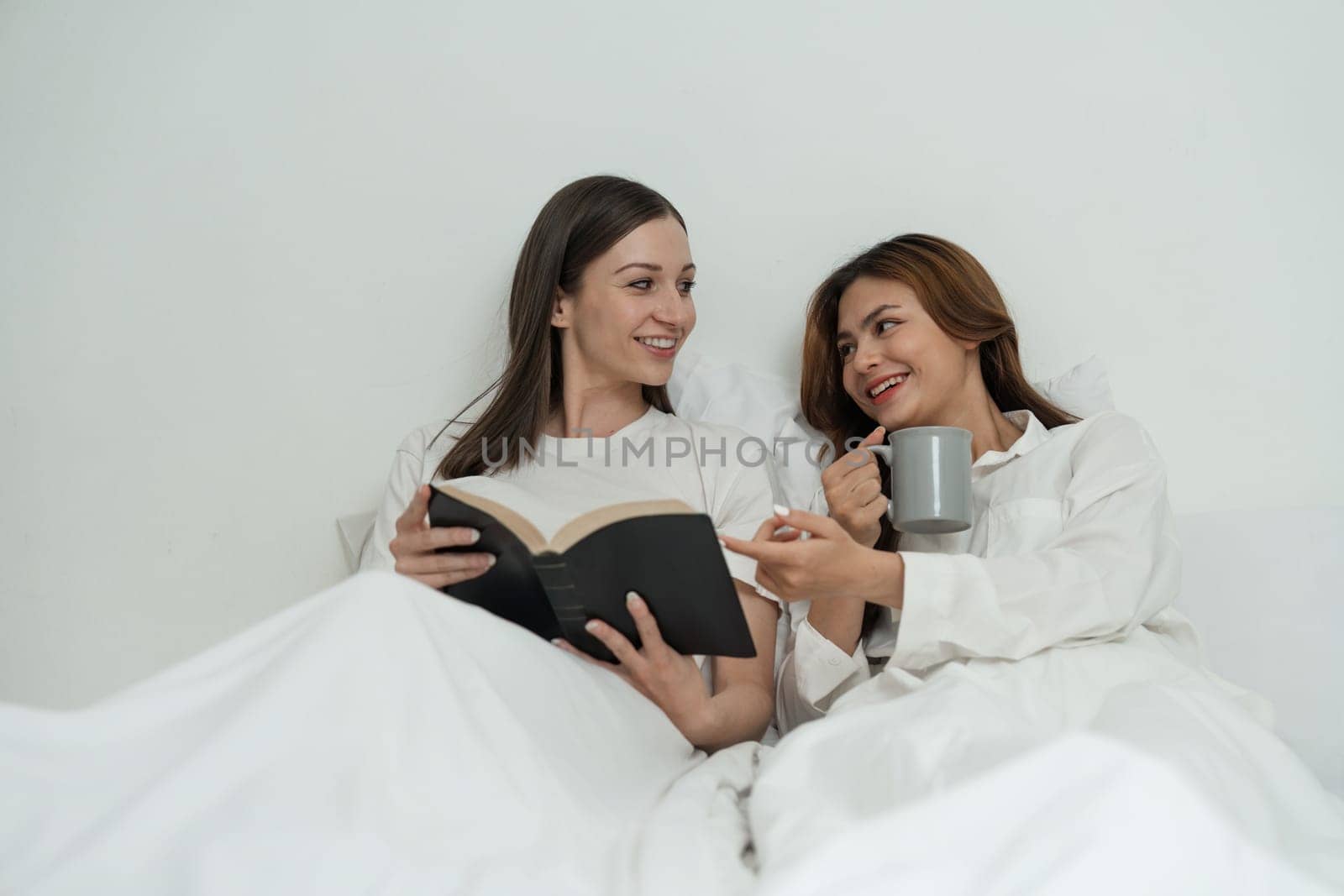 Two Asian cheerful happy female girlfriends having a coffee sitting on the bed while looking at the book. Concept lifestyle lesbian couple by nateemee