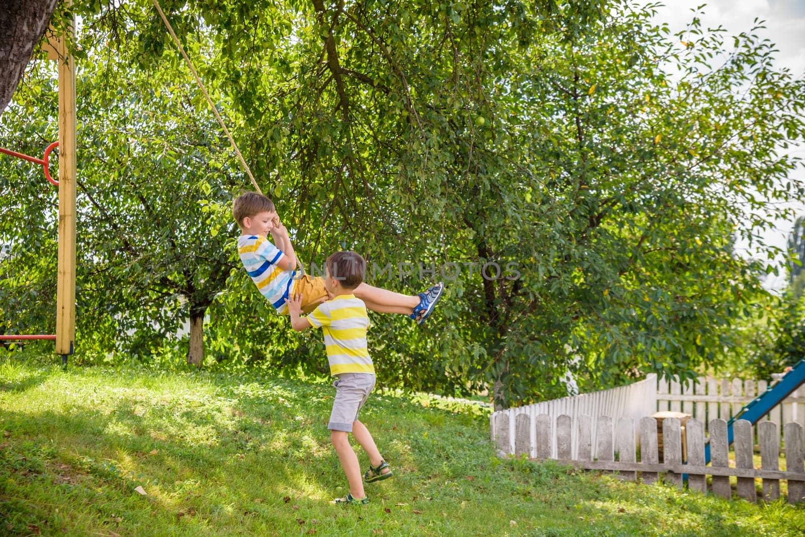 Two adorable happy little boys is having fun on a rope swing which he has found while having rest outside city. Active leisure time with children by Kobysh