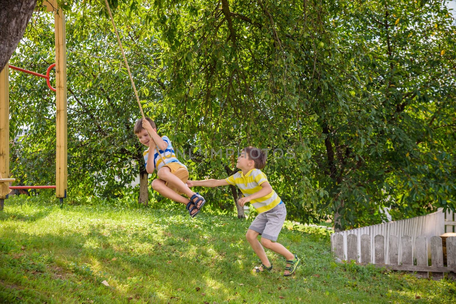 Two adorable happy little boys is having fun on a rope swing which he has found while having rest outside city. Active leisure time with children.