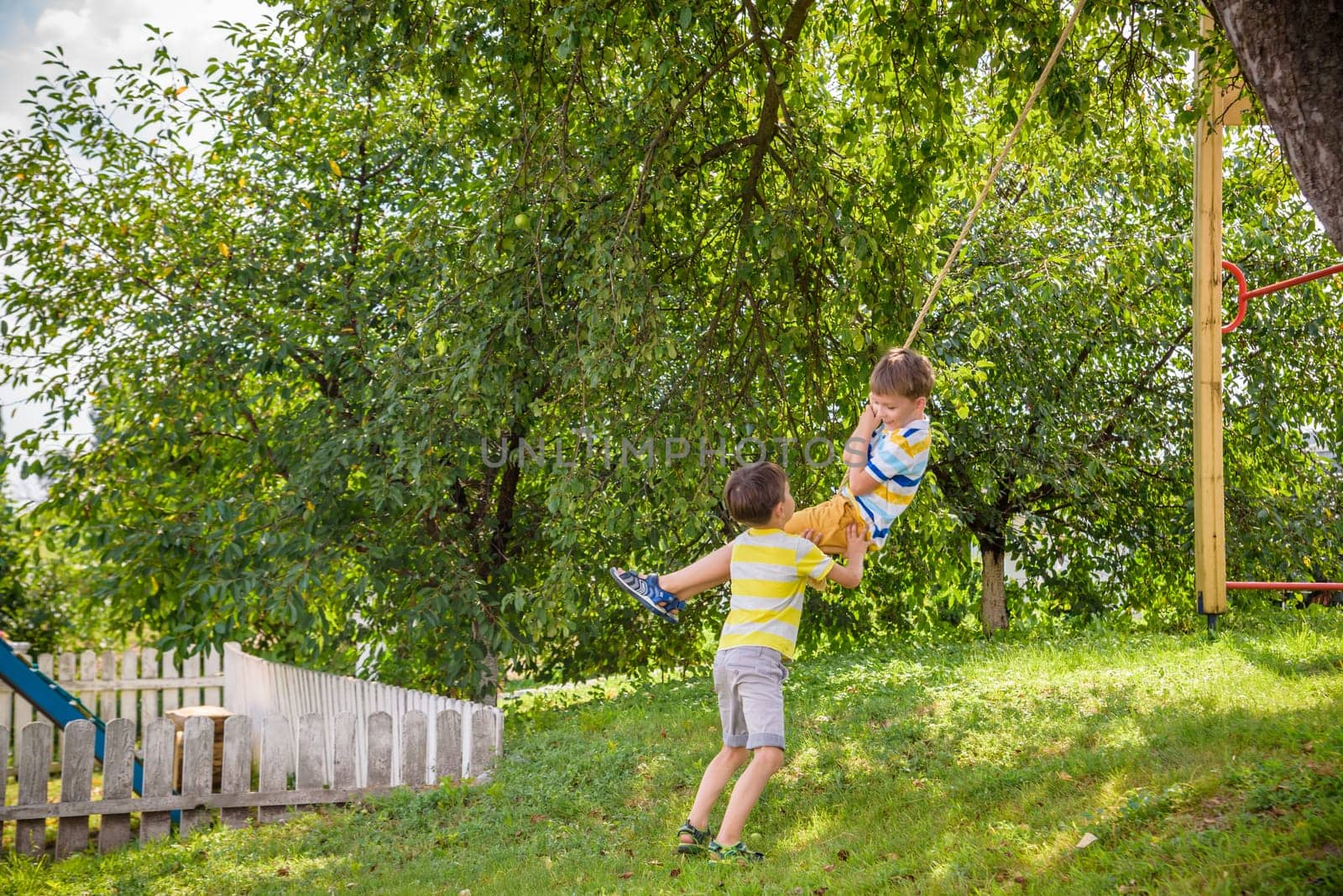Two adorable happy little boys is having fun on a rope swing which he has found while having rest outside city. Active leisure time with children.