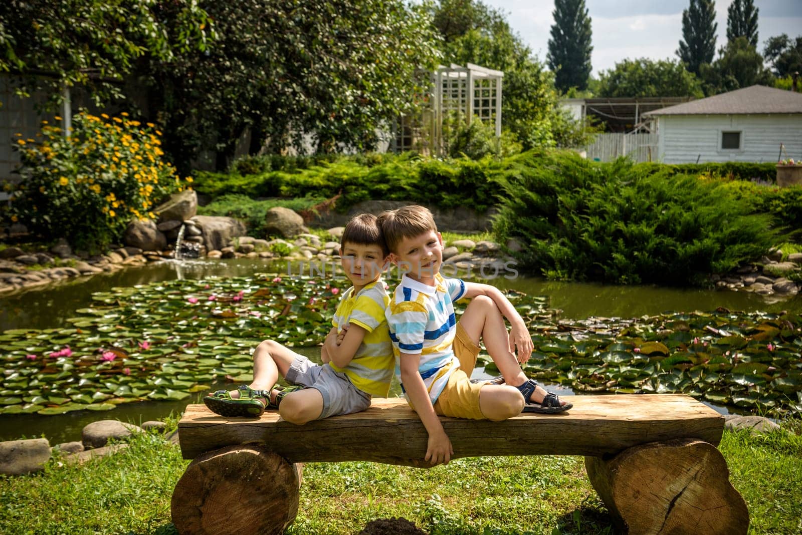 Two brother boys sitting on a bench in a park near peaceful lake with water lily Nymphaeum. Kids relaxing on nature on hot summer day.