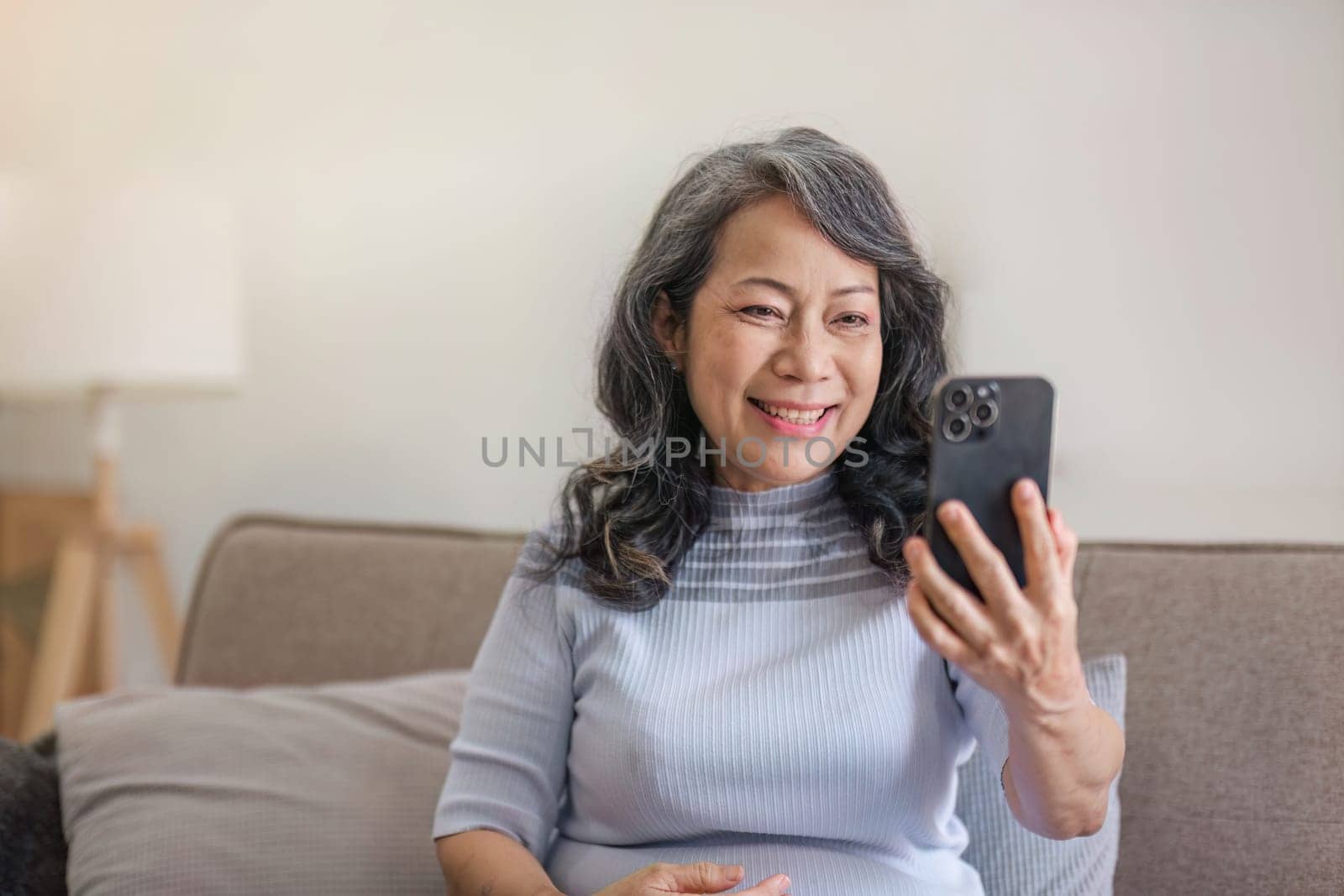 Portrait, Happy 60s retired Asian woman using her phone on a sofa in the living room. chatting, scrolling on the phone by wichayada