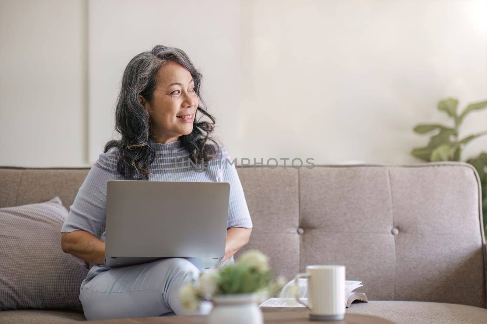 Happy Asian 60s retired woman enjoys social media on laptop while relaxing in living room..