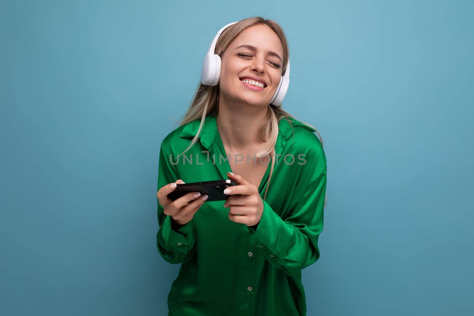 photo of a cute cheerful attractive blonde young woman in headphones with a smartphone in her hands on a blue background.