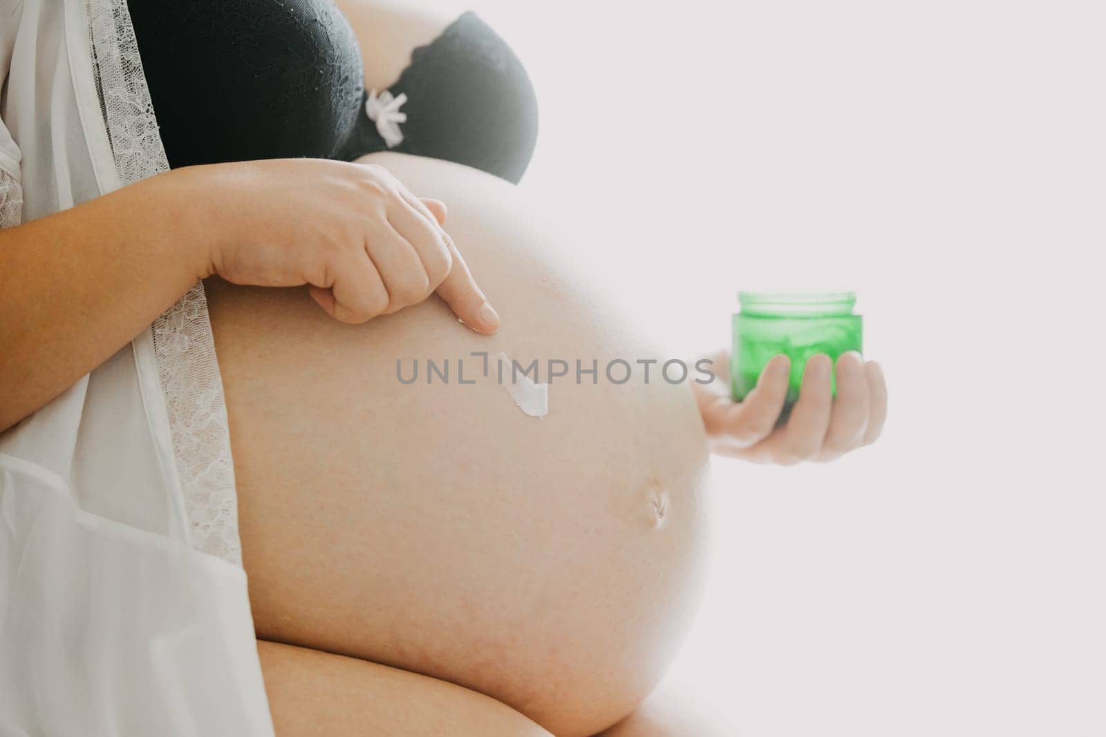 Young pregnant woman with belly smearing cream against stretch marks, pregnancy concept