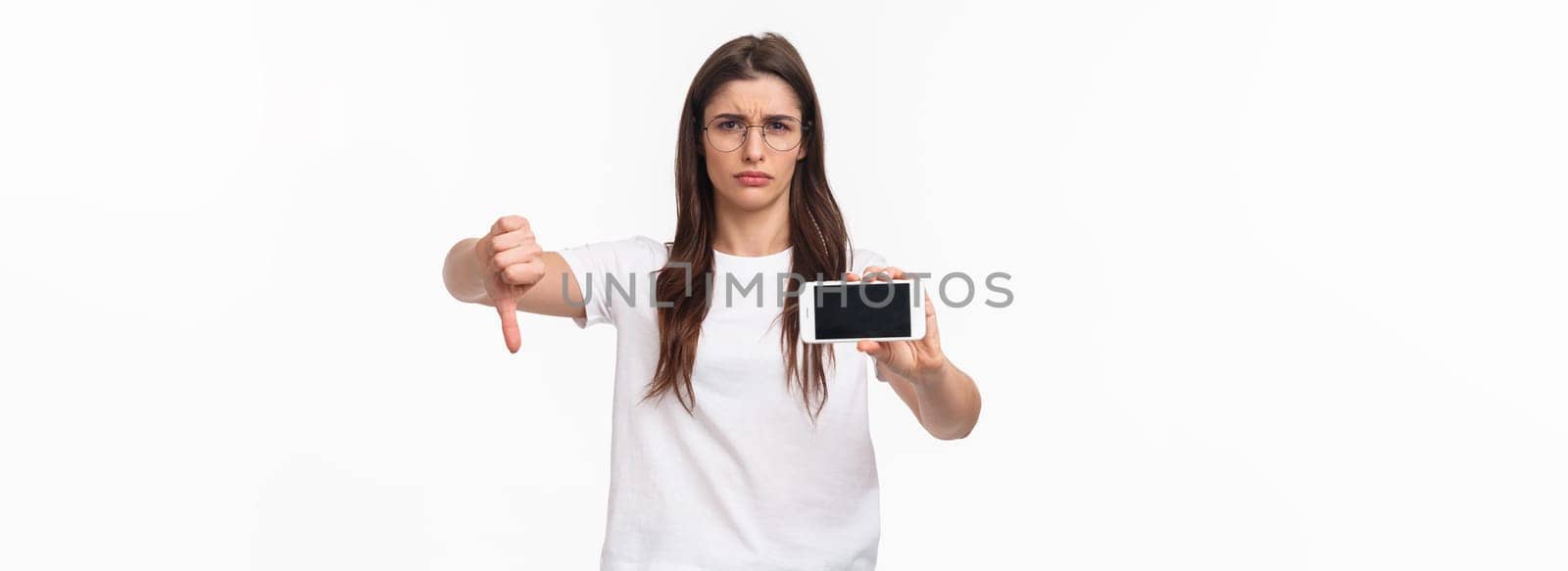 Communication, technology and lifestyle concept. Portrait of upset and disappointed young female rated bad app, showing content on mobile phone, grimacing dislike, show thumbs-down disapproval.
