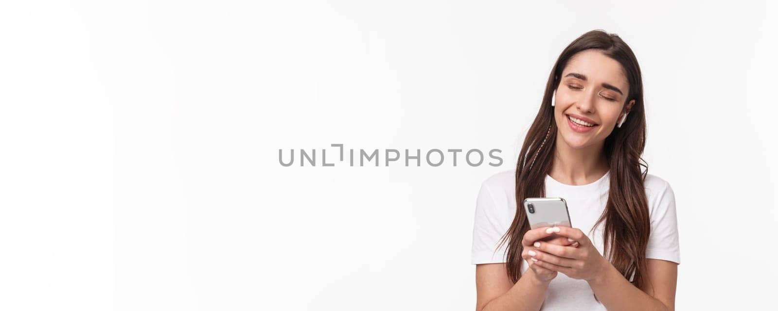 Close-up portrait of tender cute young brunette woman close eyes, shaking hand in rhythm music, smiling from delight, listening song in wireless earphones, holding smartphone by Benzoix