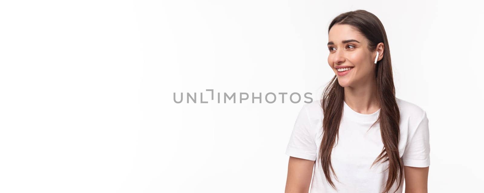 Close-up portrait of lovely, pretty young woman in t-shirt, listening music in wireless headphones, look away with pleased smile, enjoying favorite podcast or songs playlist, white background.
