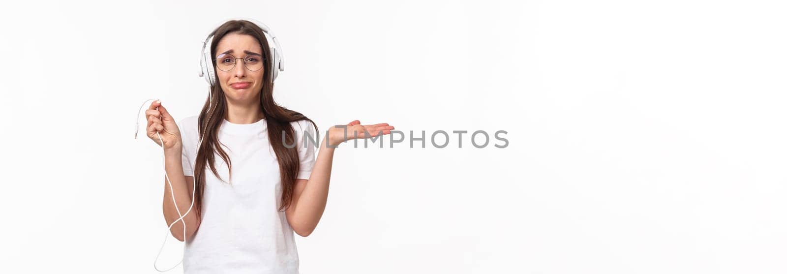 Technology, lifestyle and music concept. Portrait of awkward and frustrated young gloomy girl in headphones, holding wire and shrugging upset, earphones broke, want buy wireless earbuds.
