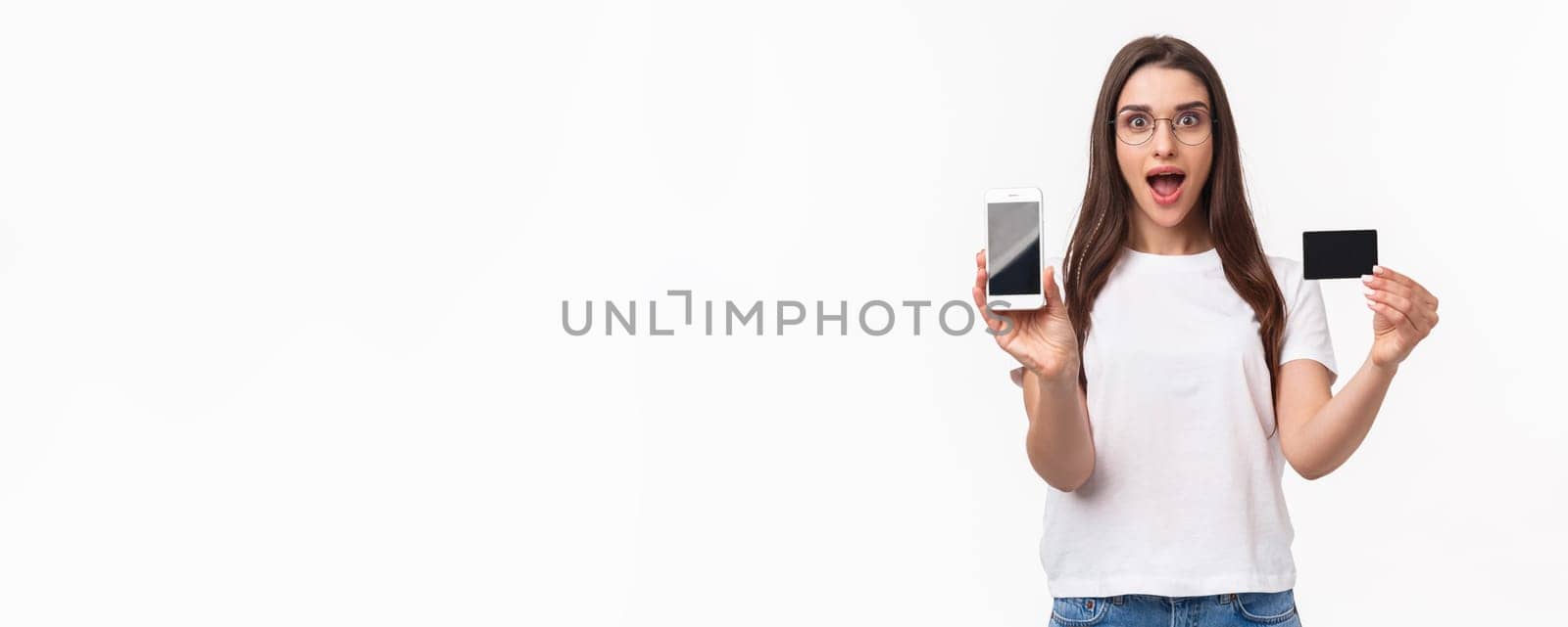 Portrait of excited and happy young woman introduce new application, favorite online store to buy clothes, show mobile phone and credit card, smiling amused, white background by Benzoix